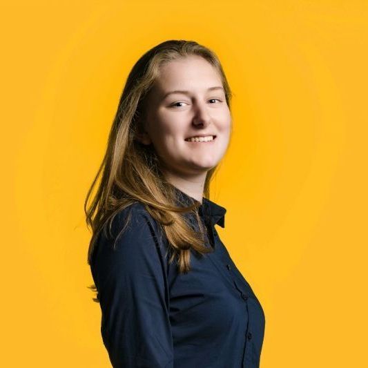 A young woman in a blue shirt is smiling in front of a yellow background.