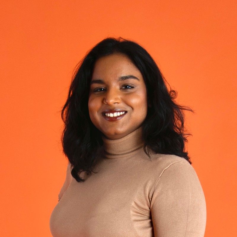 A woman wearing a tan turtleneck sweater is smiling in front of an orange background.