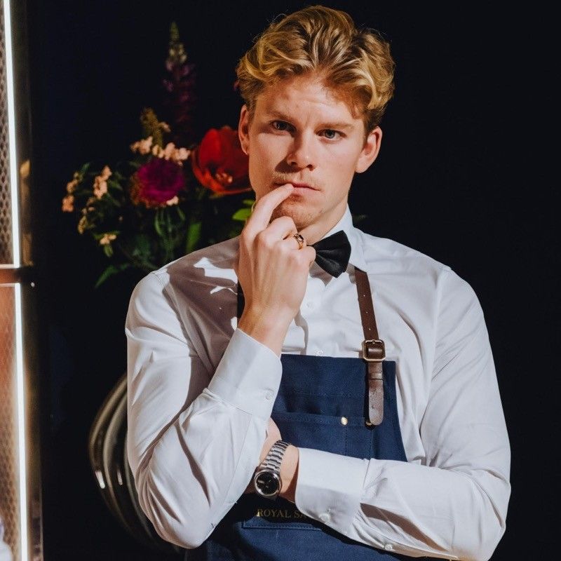 A man wearing an apron and bow tie holds his hand to his chin