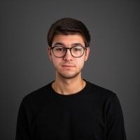 A young man wearing glasses and a black sweater is standing in front of a gray background.