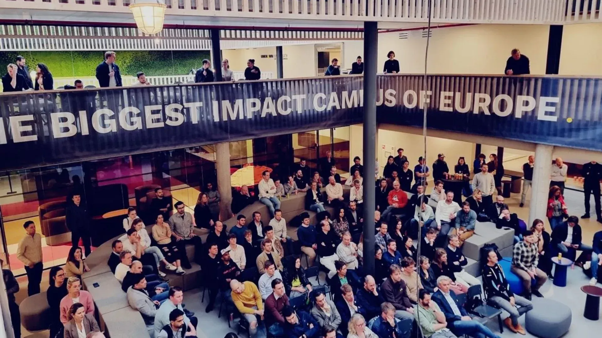 A large group of people are sitting under a sign that says the biggest impact campus of europe.