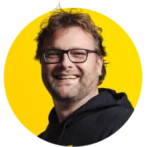 A man wearing glasses and a black hoodie is smiling in a yellow circle.