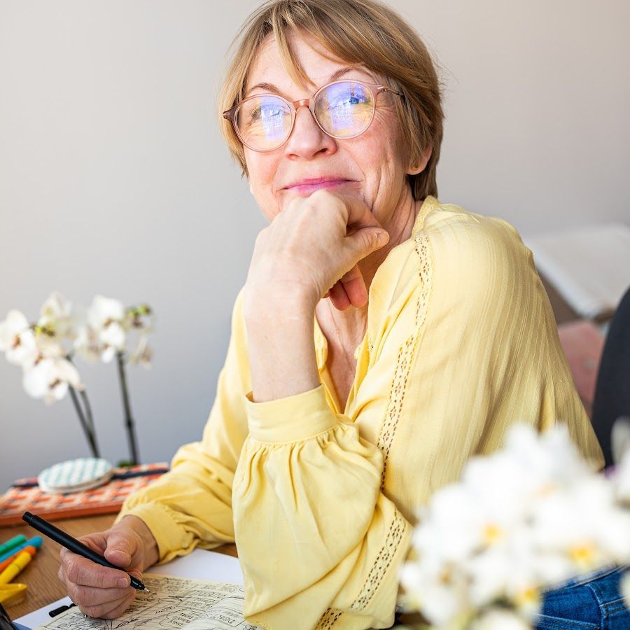 A woman wearing glasses and a yellow shirt is sitting at a table with a pen in her hand.
