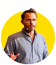 A man in a blue shirt is standing in front of a yellow circle and talking into a microphone.