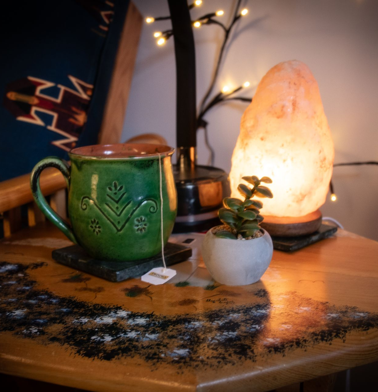 A green mug with a tea bag, a small succulent, and a glowing salt lamp sit on a decorative wooden table with fairy lights.