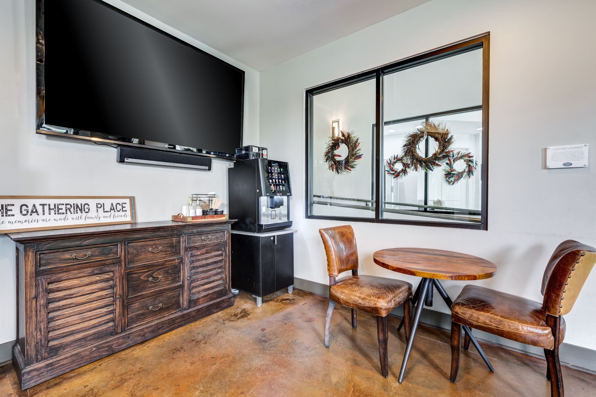 A cozy lounge area with TV, coffee machine, table, chairs, and large window with wreaths at Town Center at Lake Carolina in Columbia, SC.