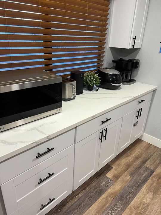 A kitchen with white cabinets , a microwave , a coffee maker , and a window.