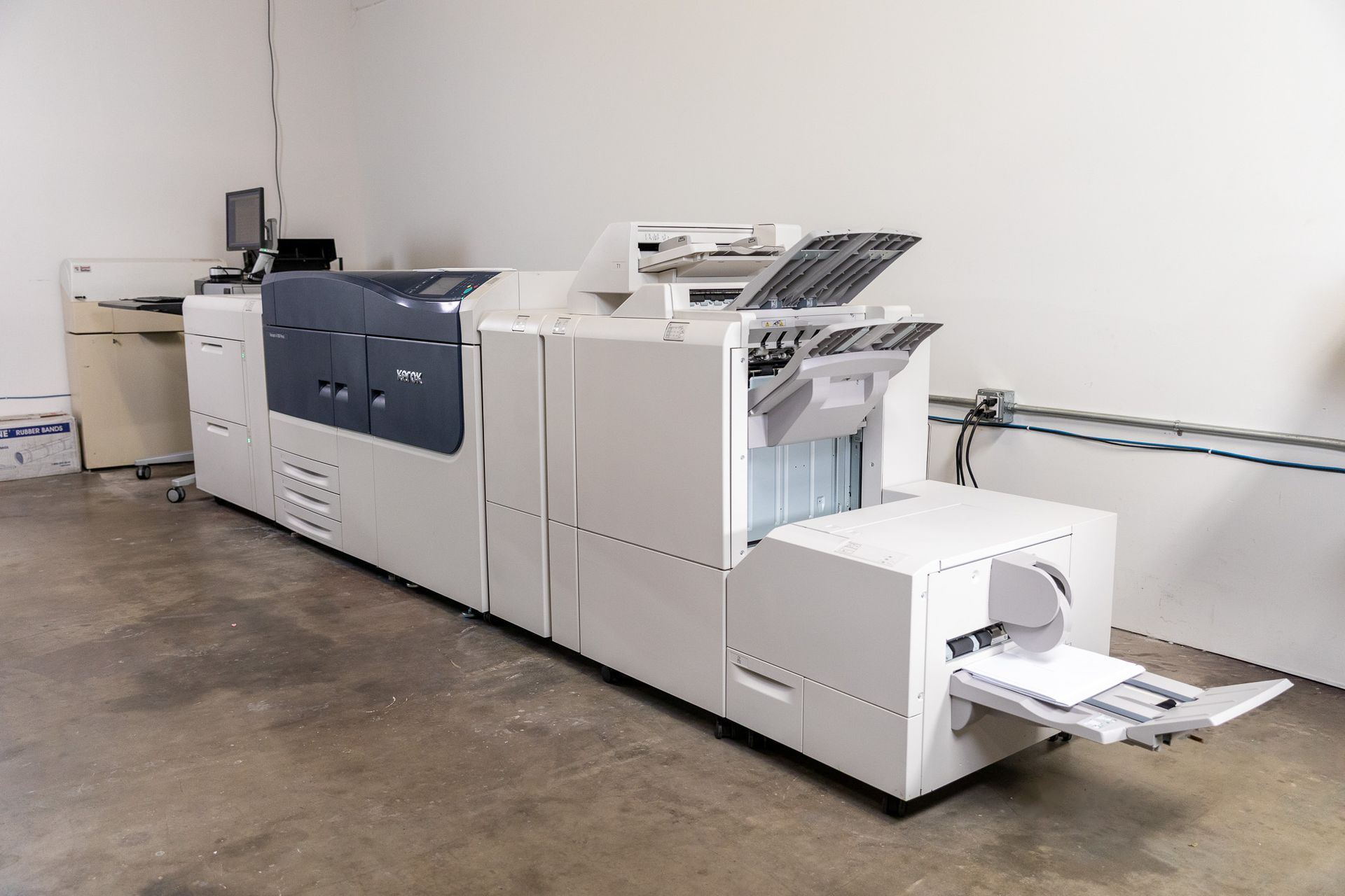 Large commercial printer in a white room, with paper output tray and control panel.