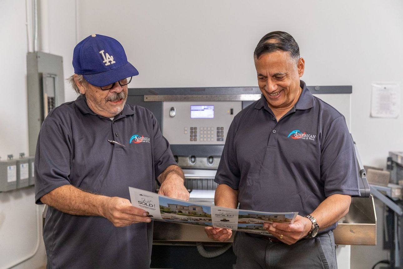 Two men reviewing printed materials in a print shop. One wears a blue cap. Both smile. Grey shirts.