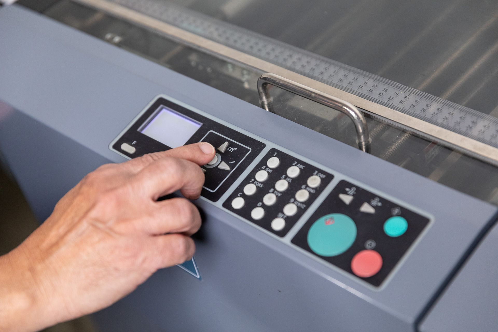 Hand pressing buttons on the control panel of a gray industrial machine with a ruler on top.