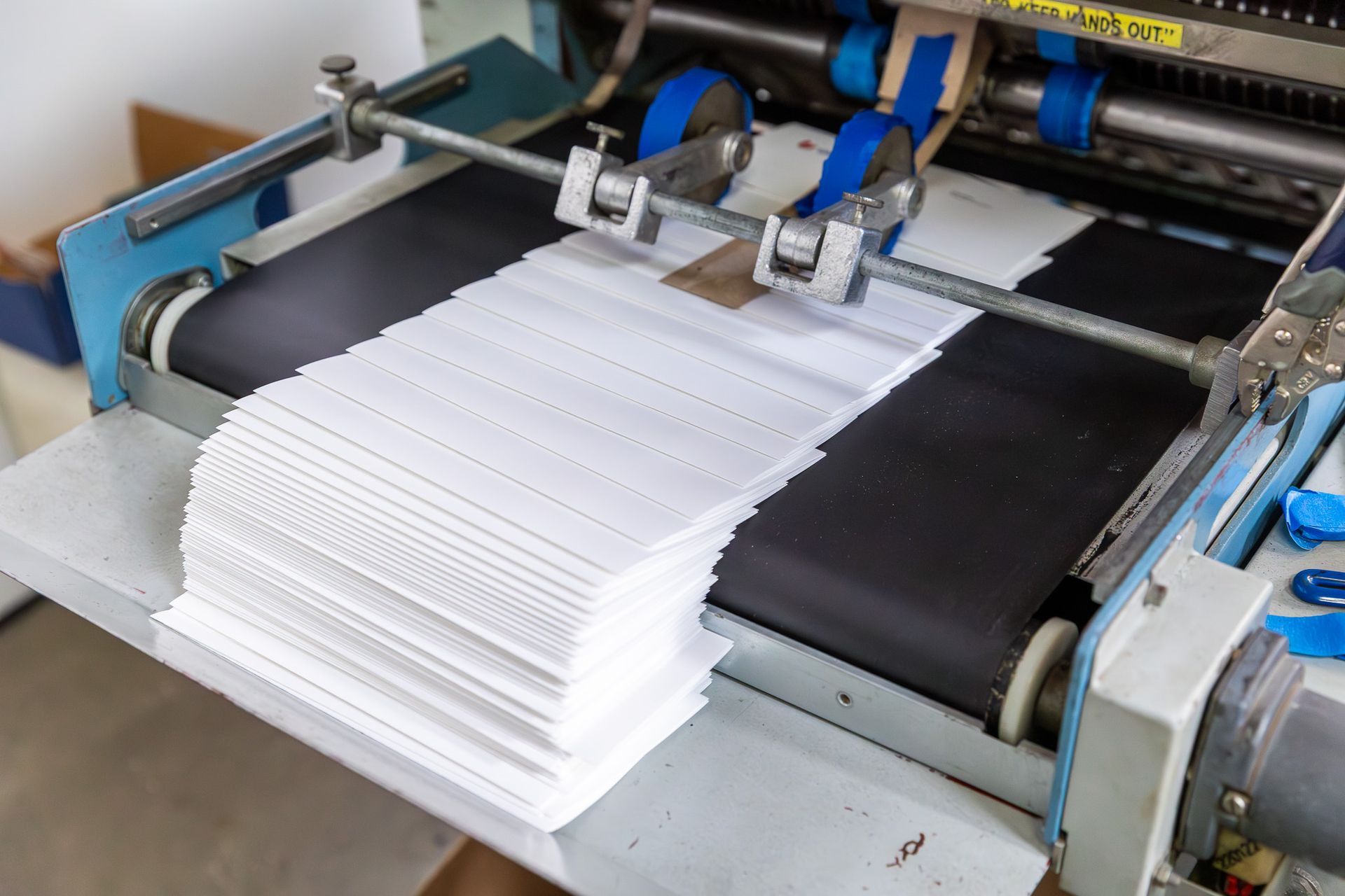 White paper stack feeding into an automated machine with rollers and a black conveyor belt.