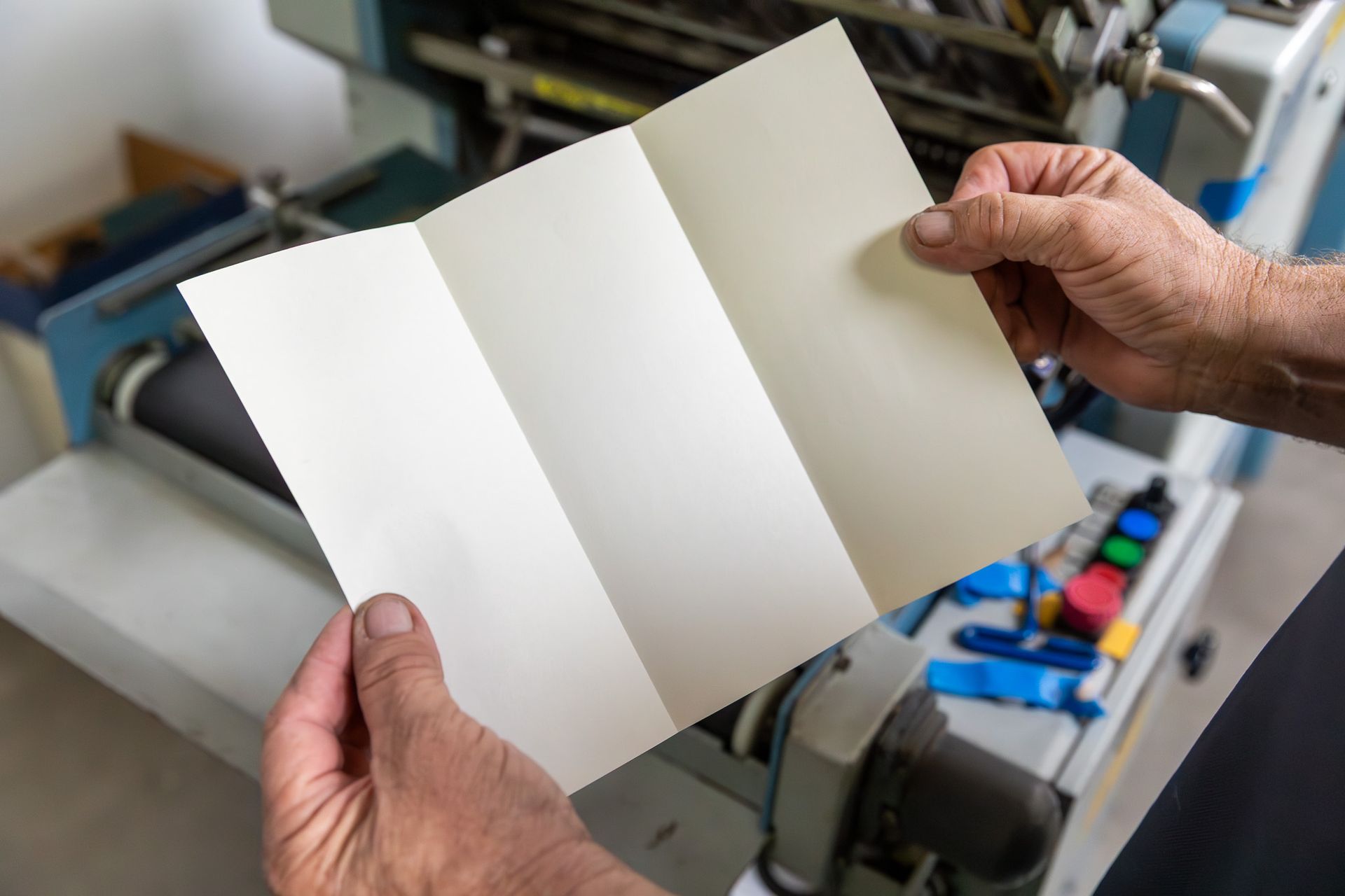 Person holding a tri-fold brochure, likely freshly printed, near a printing press.