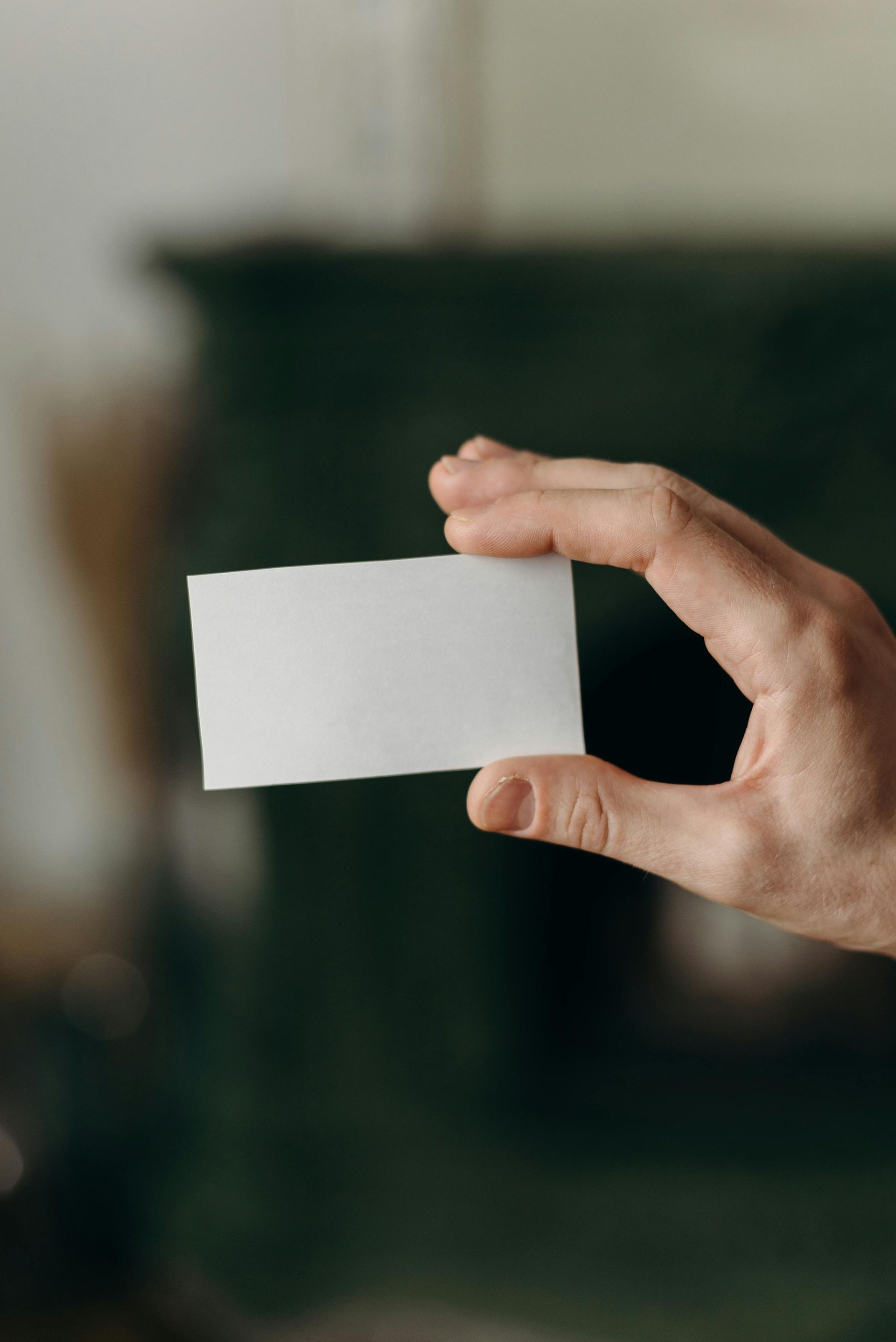 Hand holding a blank, white rectangular card against a blurred green backdrop.