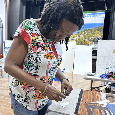 Woman cutting paper with scissors at a table, wearing a patterned shirt. Indoor setting with art materials.