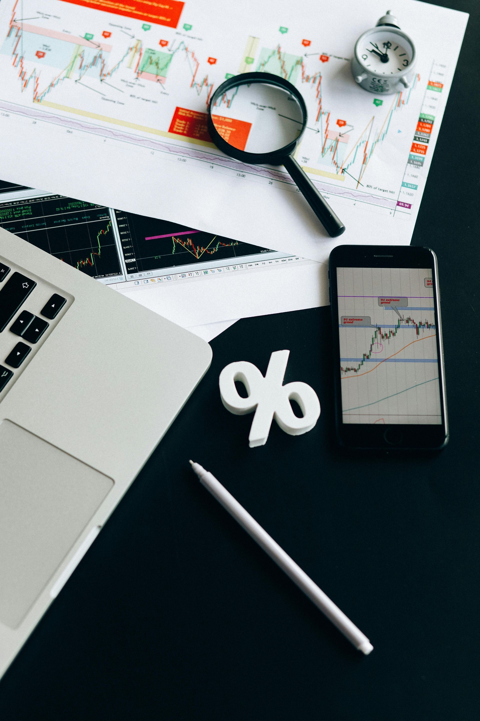 Laptop, phone, charts, magnifying glass, and percentage symbol on a dark surface, suggesting financial analysis.