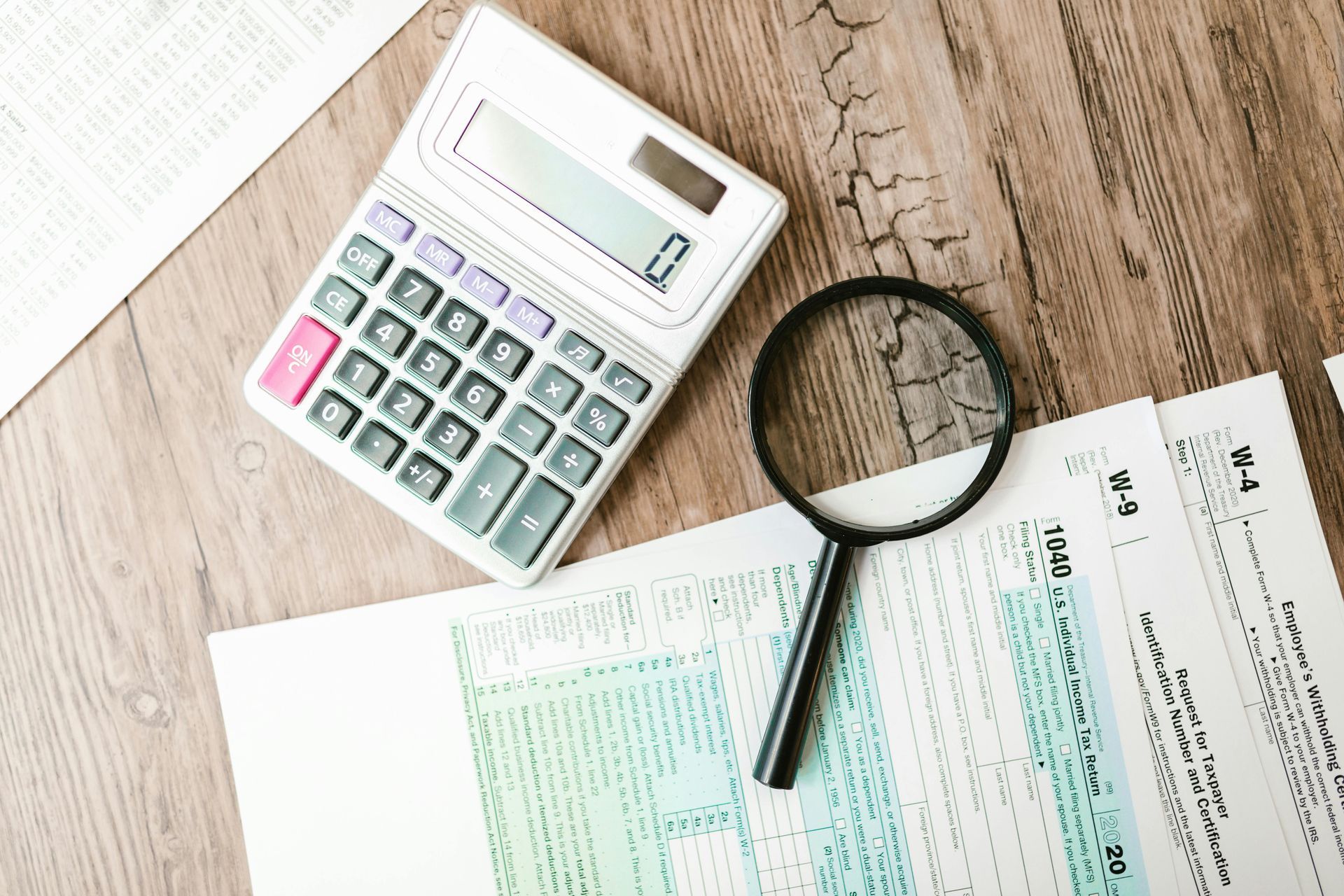 Calculator, magnifying glass, and tax forms on a wooden desk.