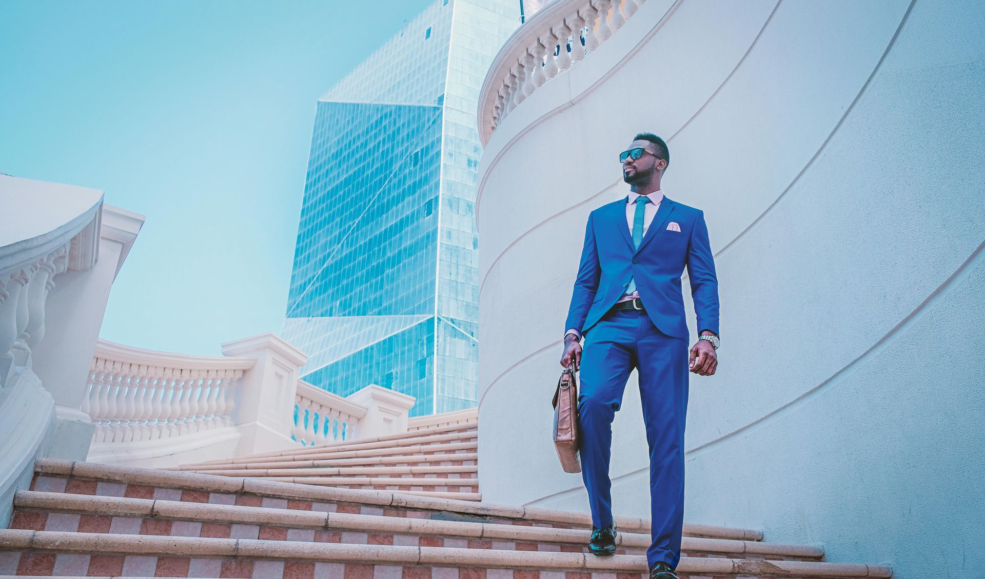 Man in blue suit and sunglasses walks down outdoor stairs, holding briefcase; modern building in background.