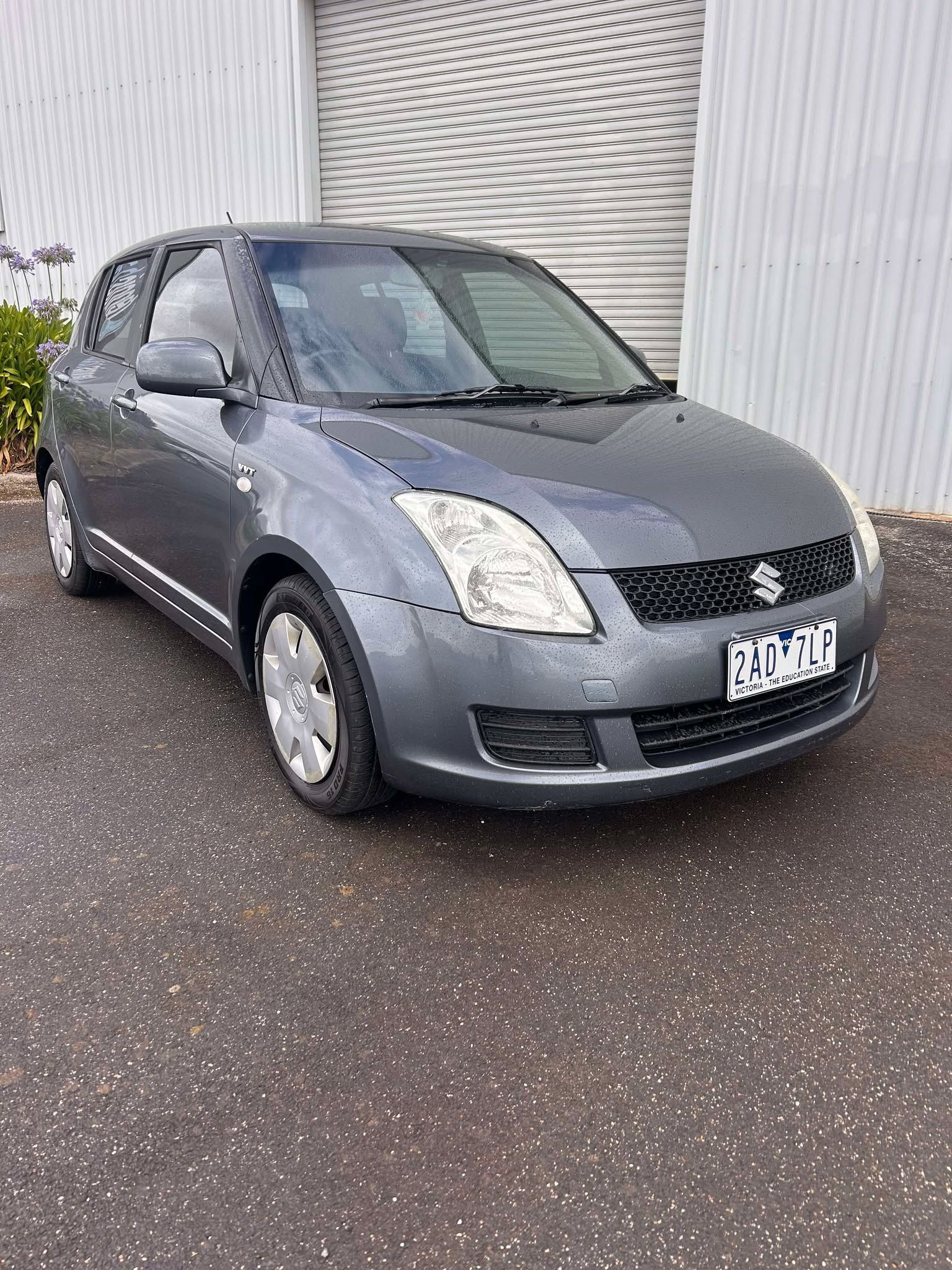 A gray suzuki swift is parked in front of a garage door.