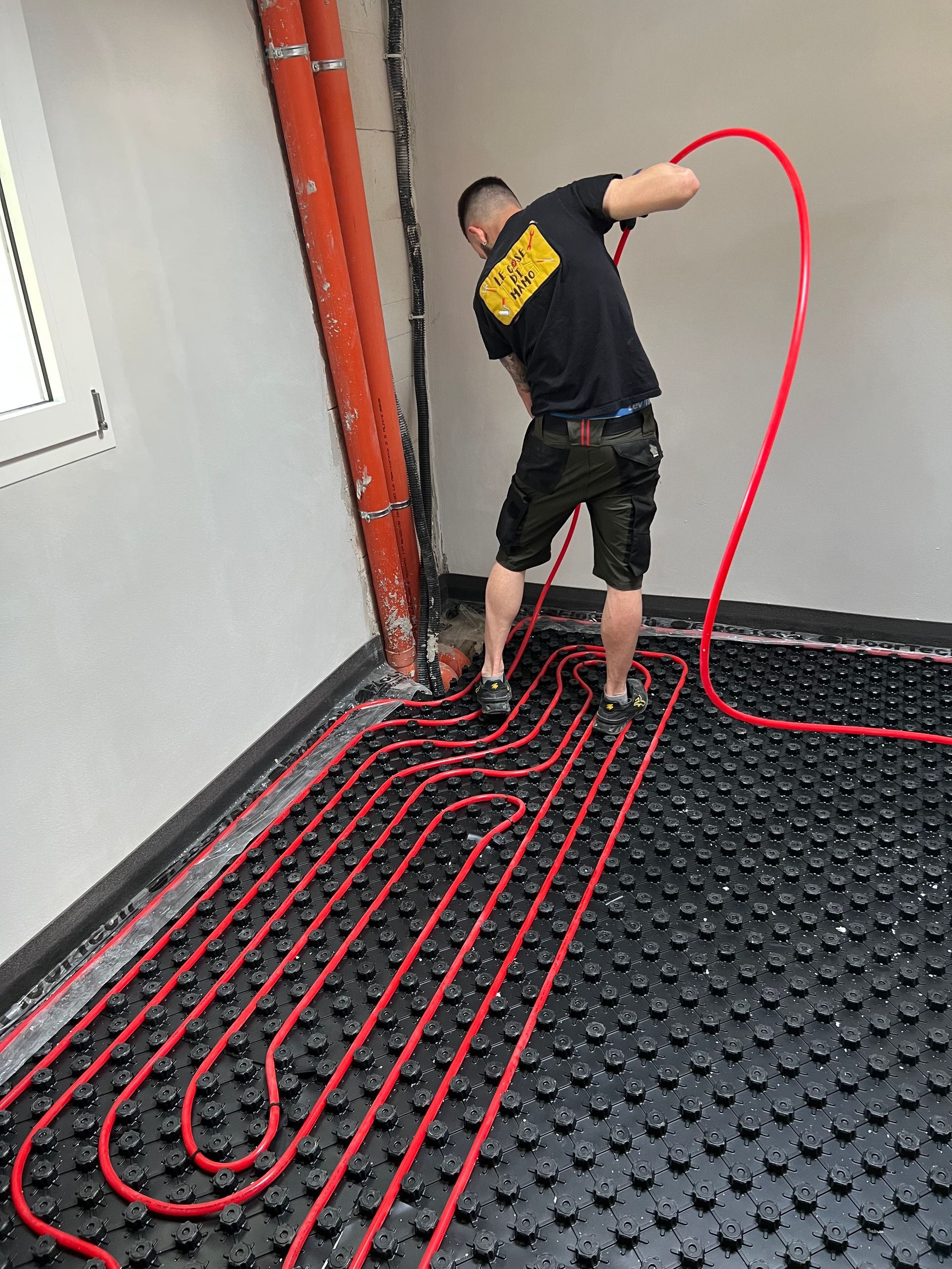 Uomo che installa tubi rossi per il riscaldamento a pavimento in una stanza. Tubi posati su un tappetino nero.