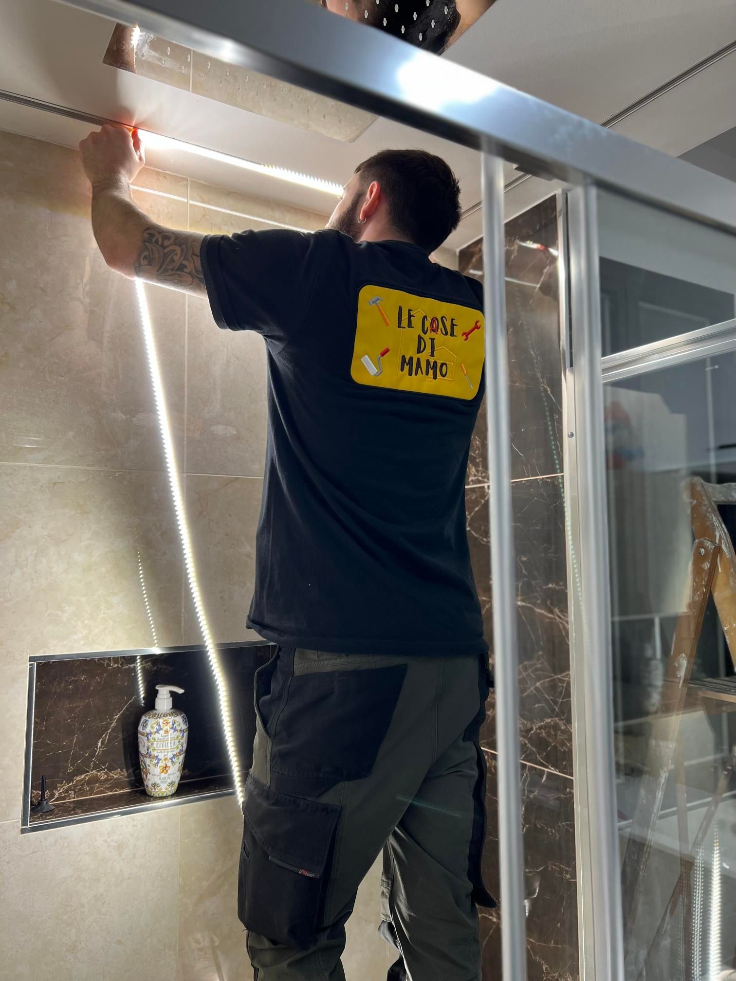 Uomo che installa il telaio della porta della doccia. Indossa una camicia nera, pantaloni scuri e lavora in un bagno.