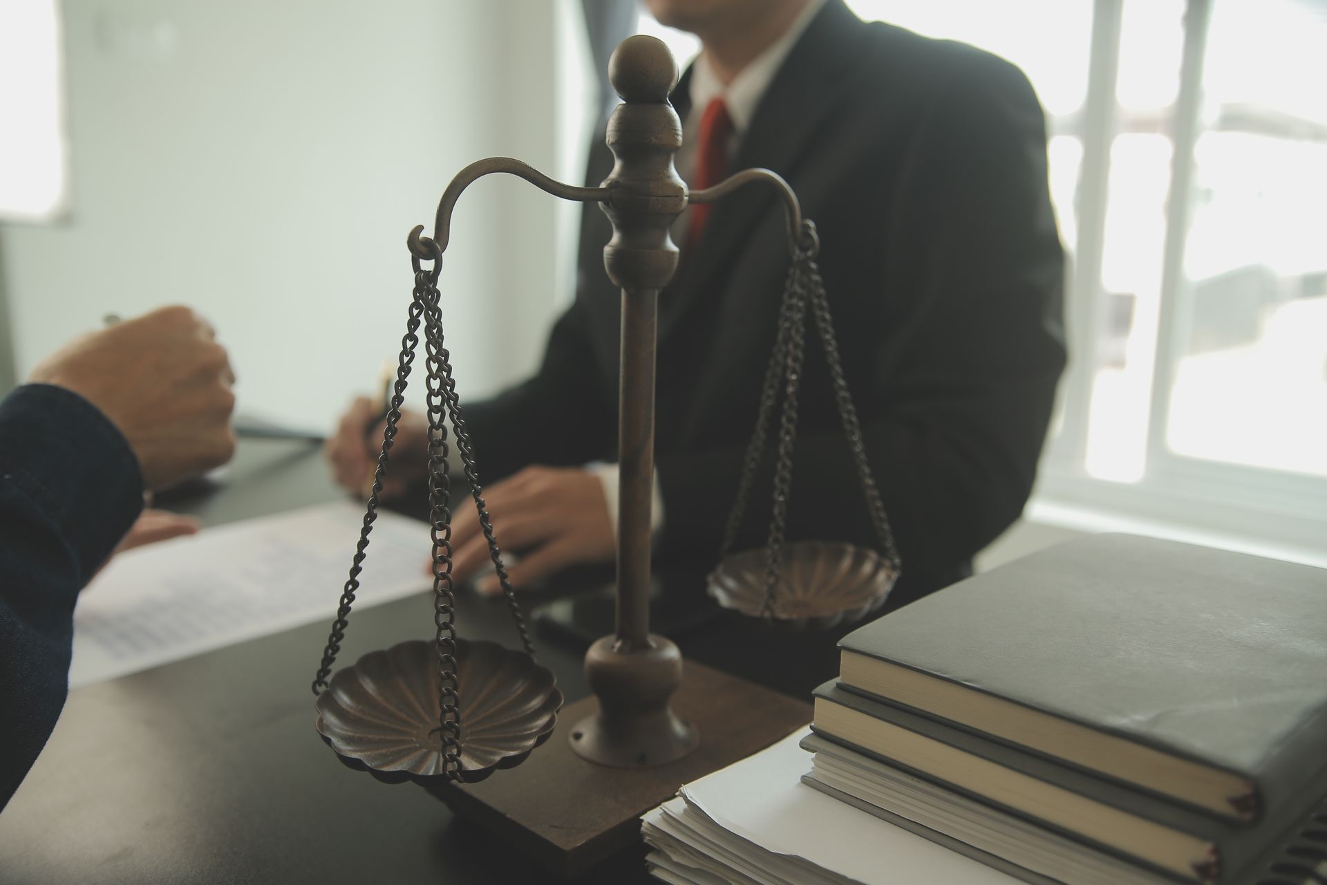 Scales of justice on a desk; two people in suits, one signing documents near window.
