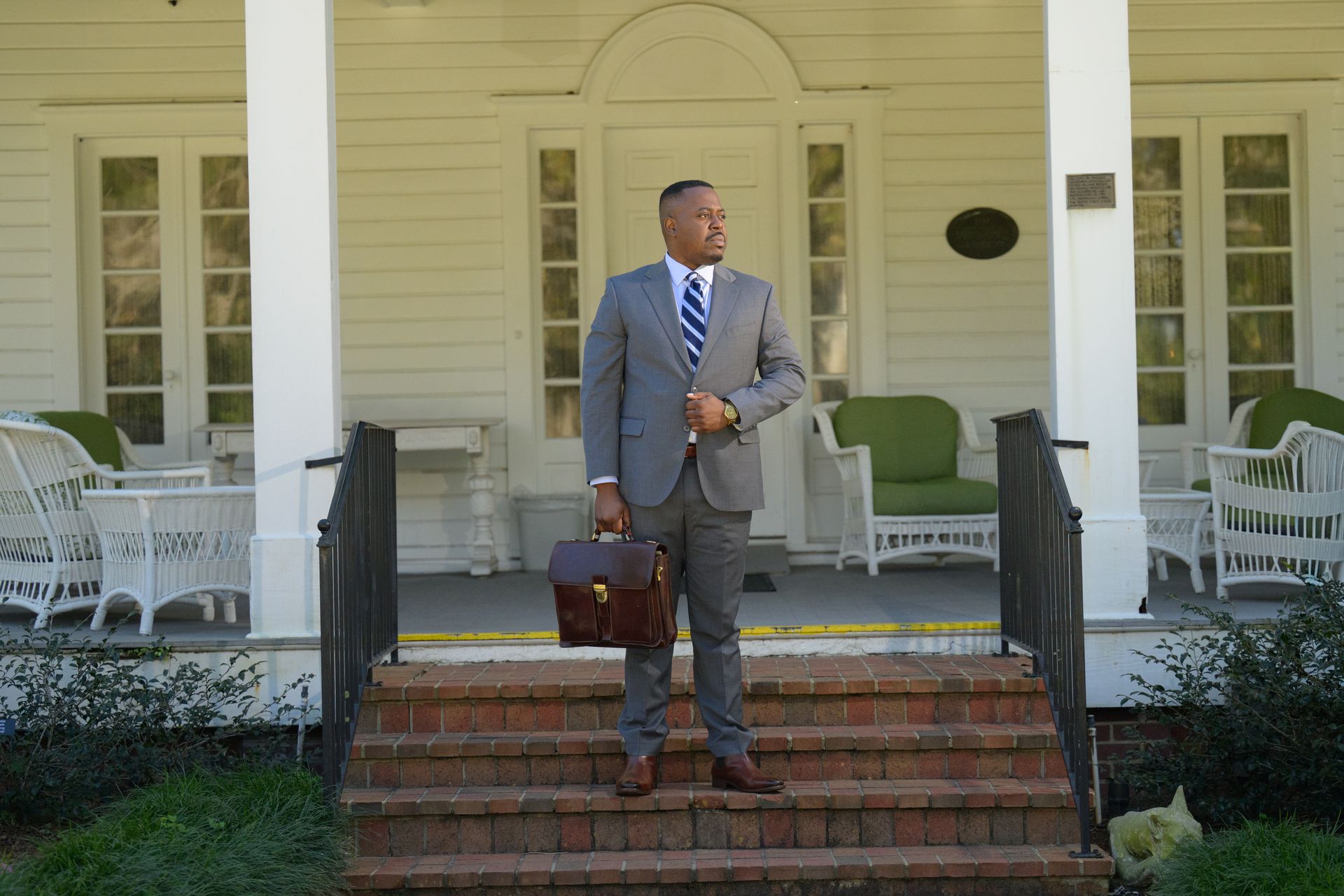 Man in a blue suit smiles, writing at a desk with a keyboard, American flag in background.