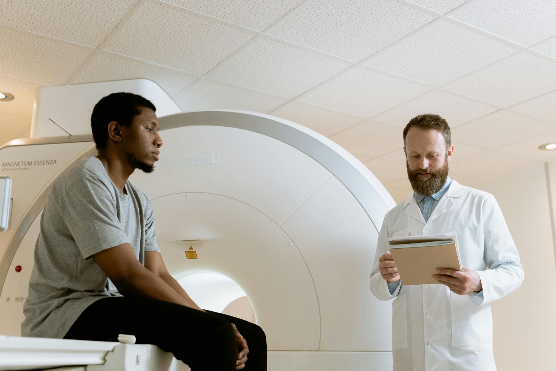 Man sits near MRI machine, doctor holds clipboard. Bright, clinical room.