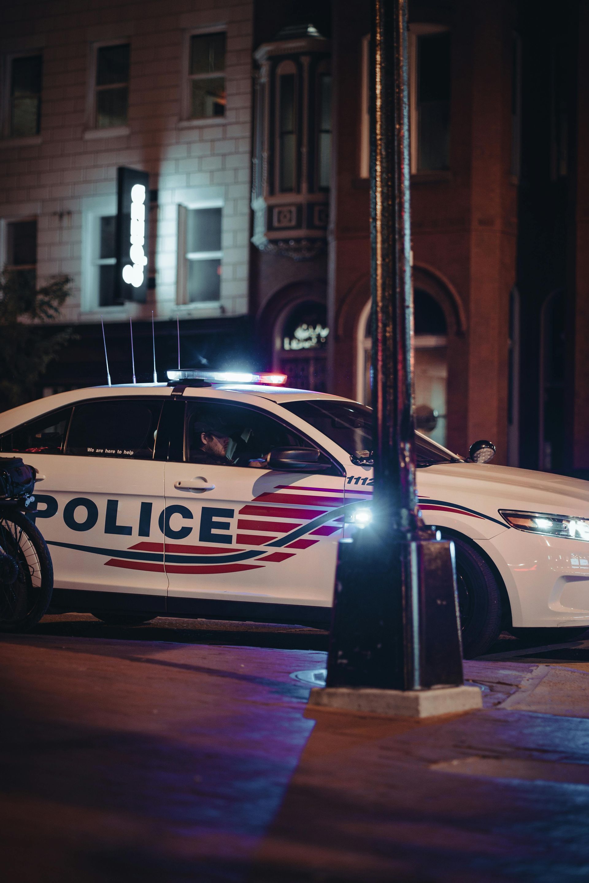 Police car parked on a city street at night with flashing lights; buildings in the background.