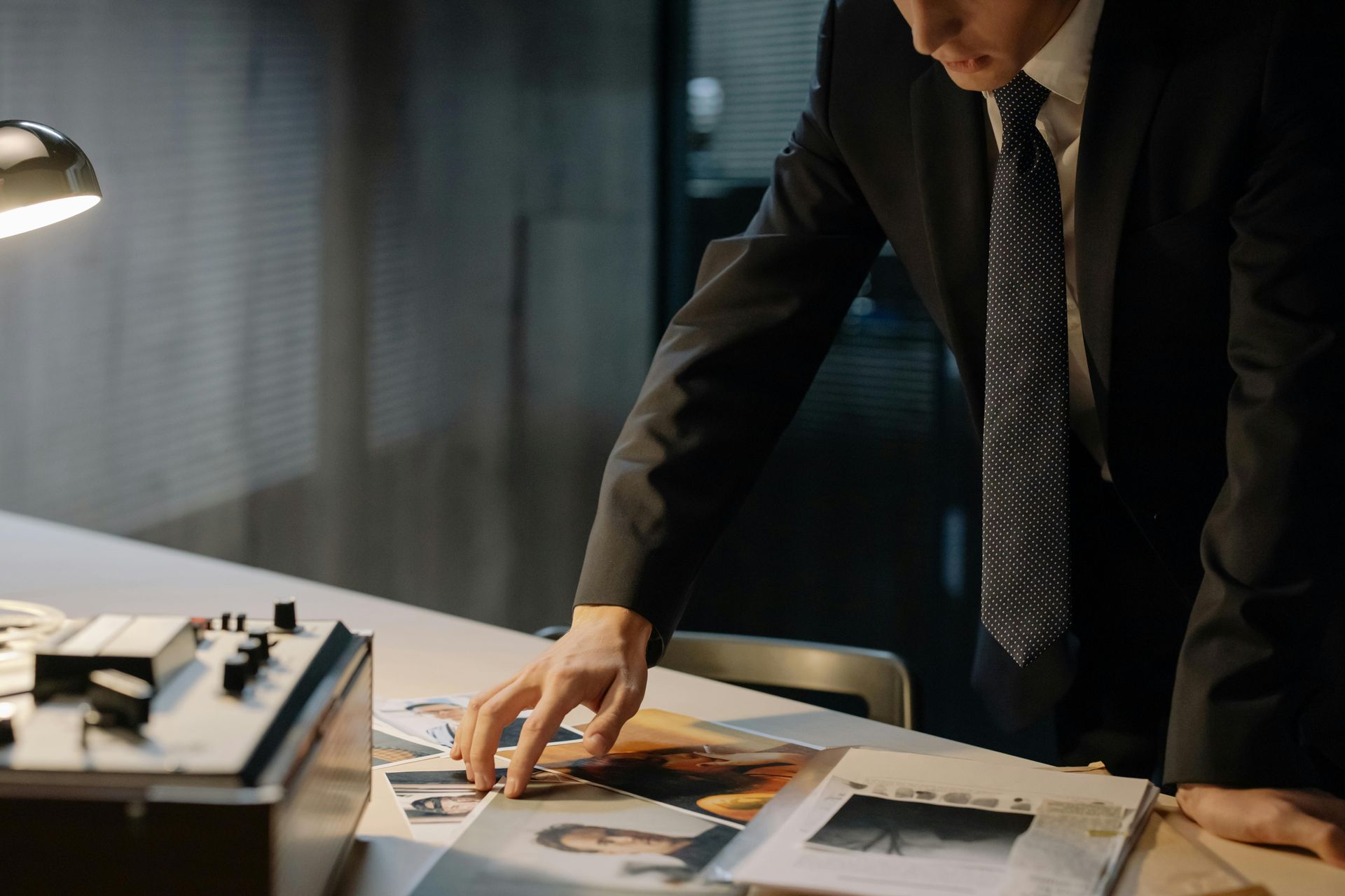 Man in suit examines photos on a desk under a desk lamp.