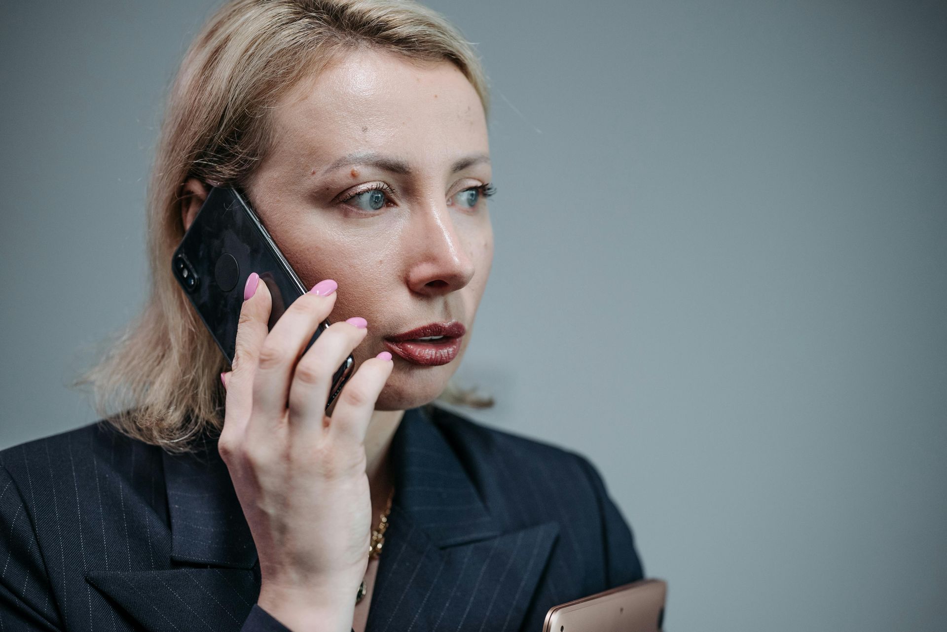 Woman in navy blazer talks on phone, looking concerned with perspiration on her face.