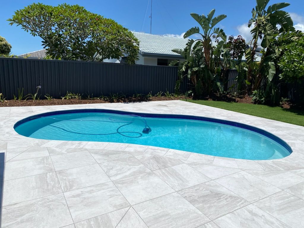 Oval-shaped swimming pool in a paved backyard, surrounded by green grass and a gray fence.