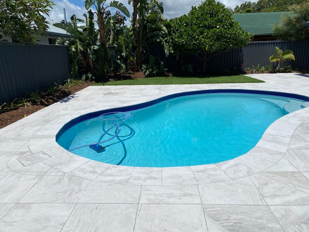 Swimming pool with blue water and light gray patio tiles, surrounded by green grass and plants.