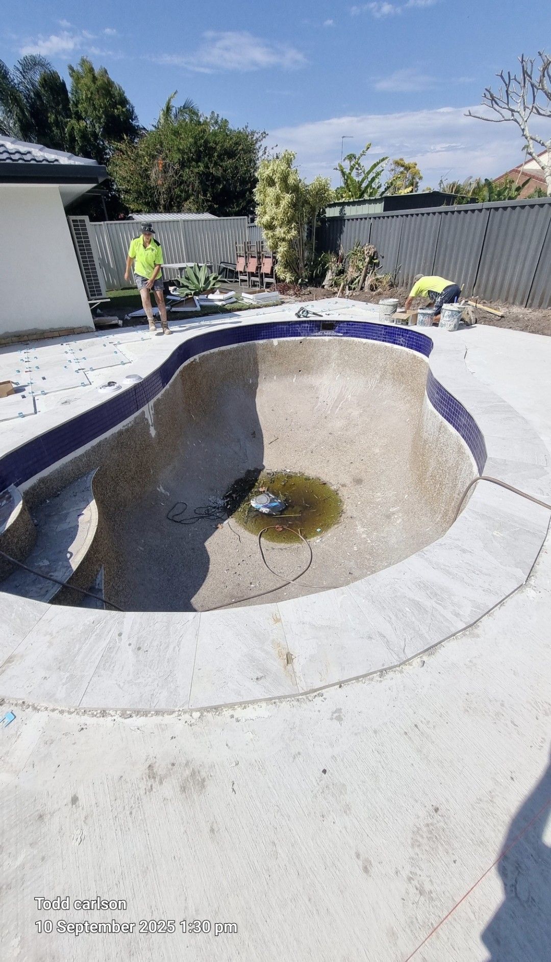 A drained swimming pool with workers, concrete, and surrounding yard under a bright sky.