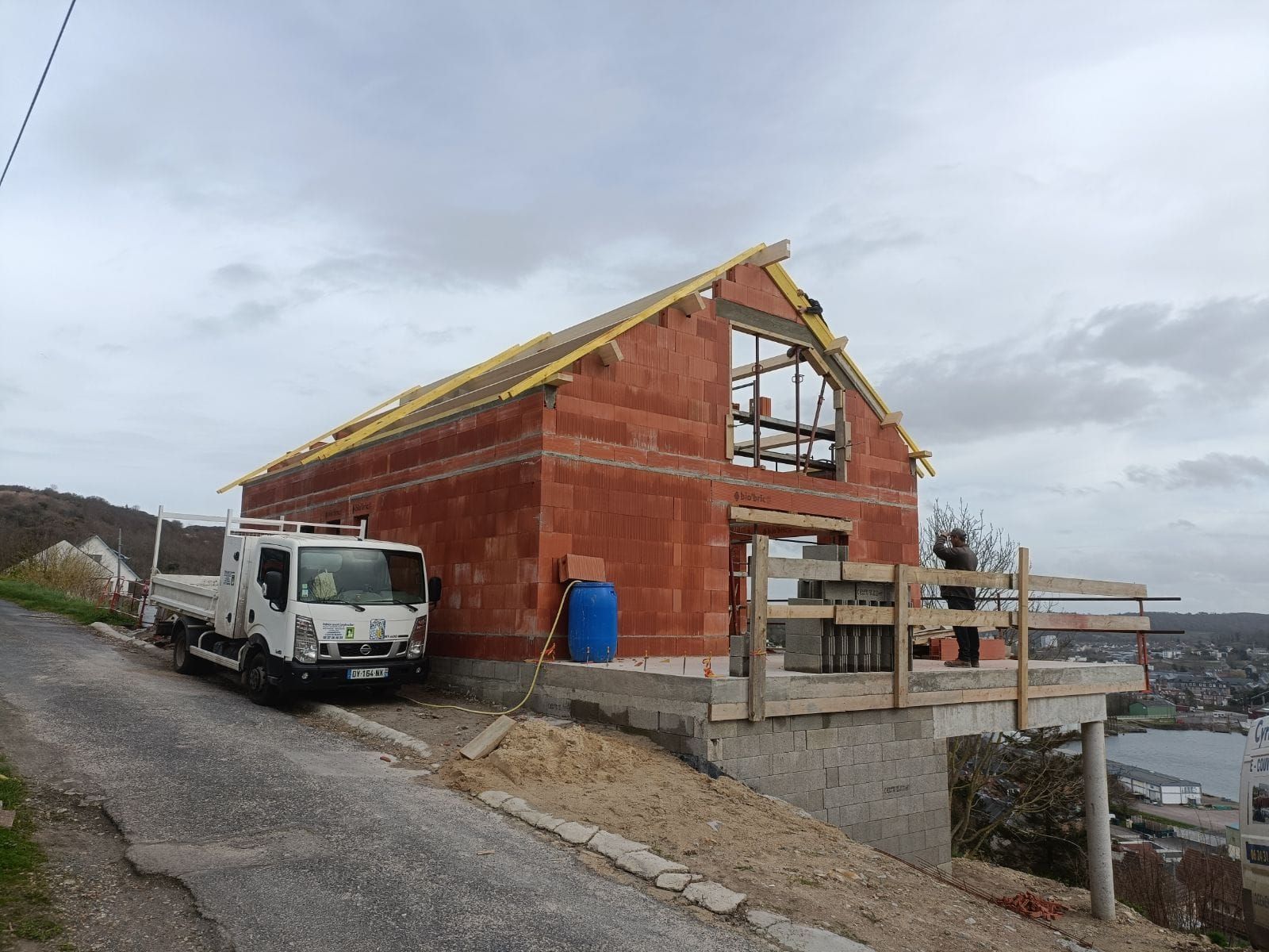 Bâtiment en construction, façade en briques rouges, avec un camion garé sur une route en pente, surplombant un plan d'eau.