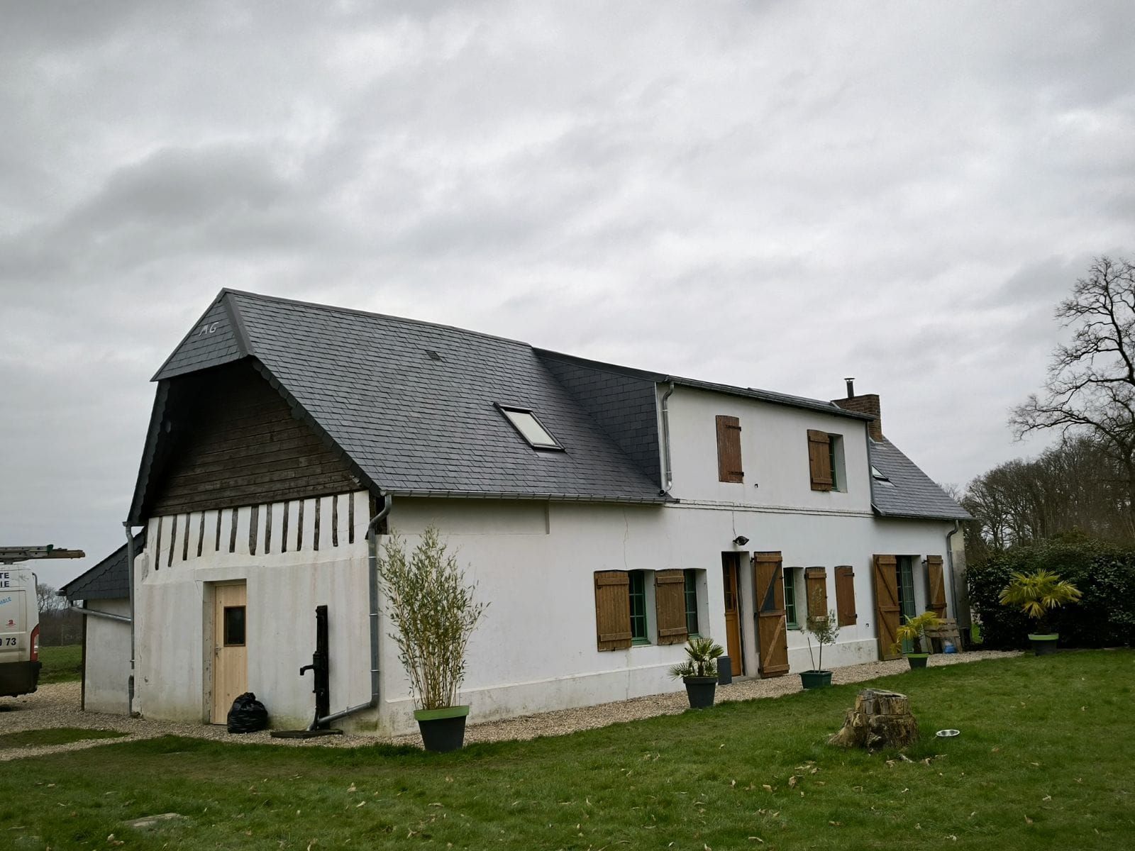 Maison blanche au toit gris foncé et aux volets marron sous un ciel nuageux.