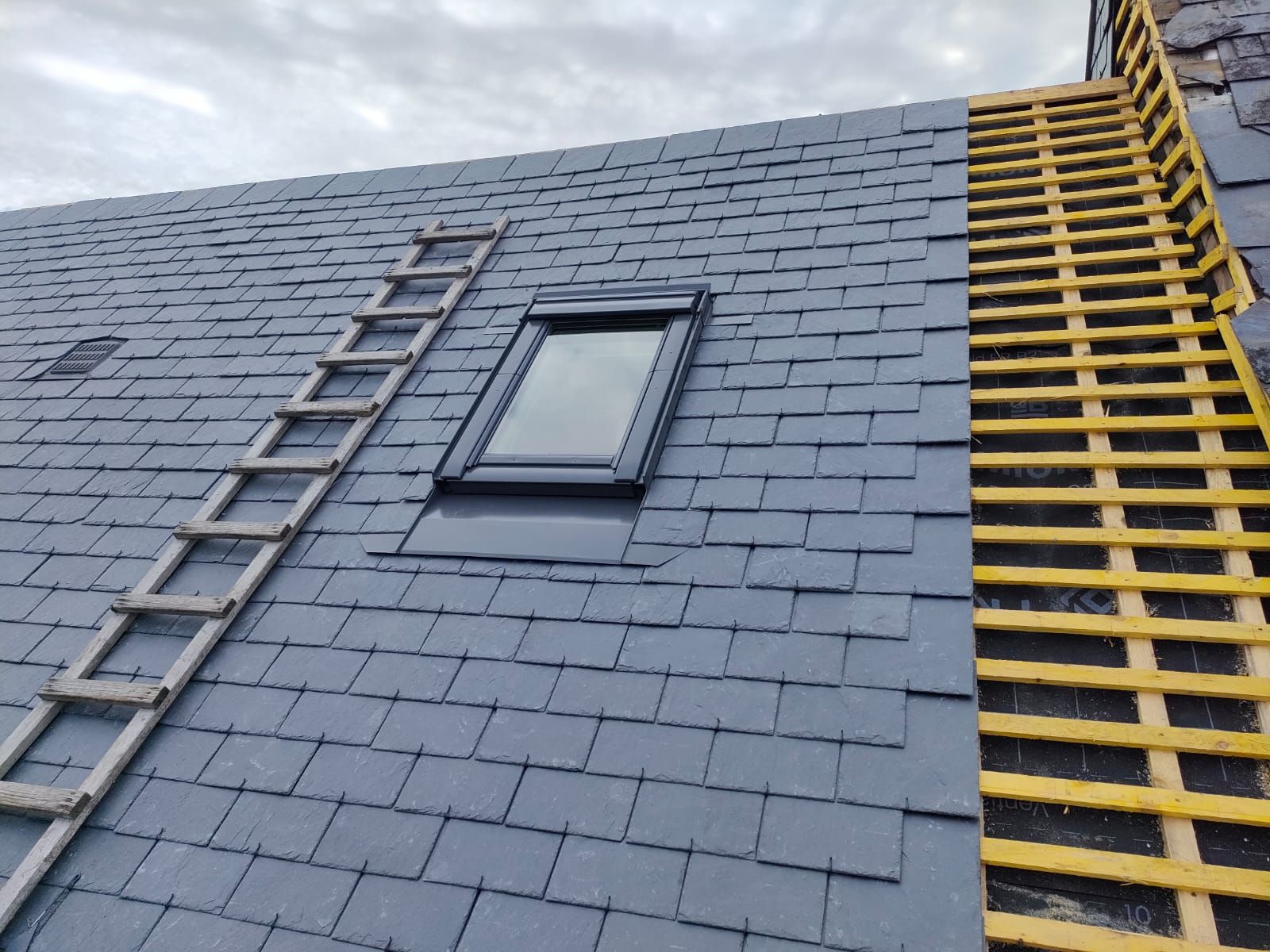Un toit en ardoise en construction ; on aperçoit un puits de lumière, une échelle et les supports de toit apparents.