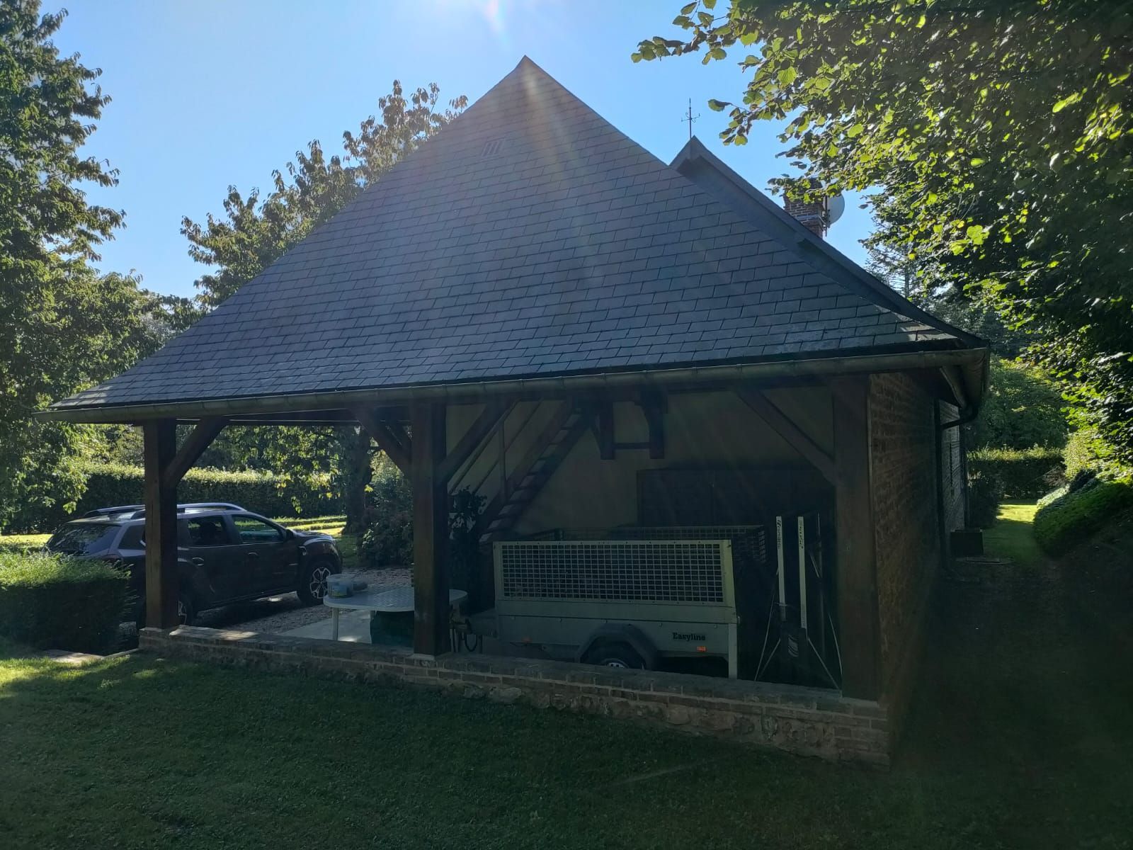 Un abri de voiture en bois au toit gris, des voitures garées et une remorque sous l'abri