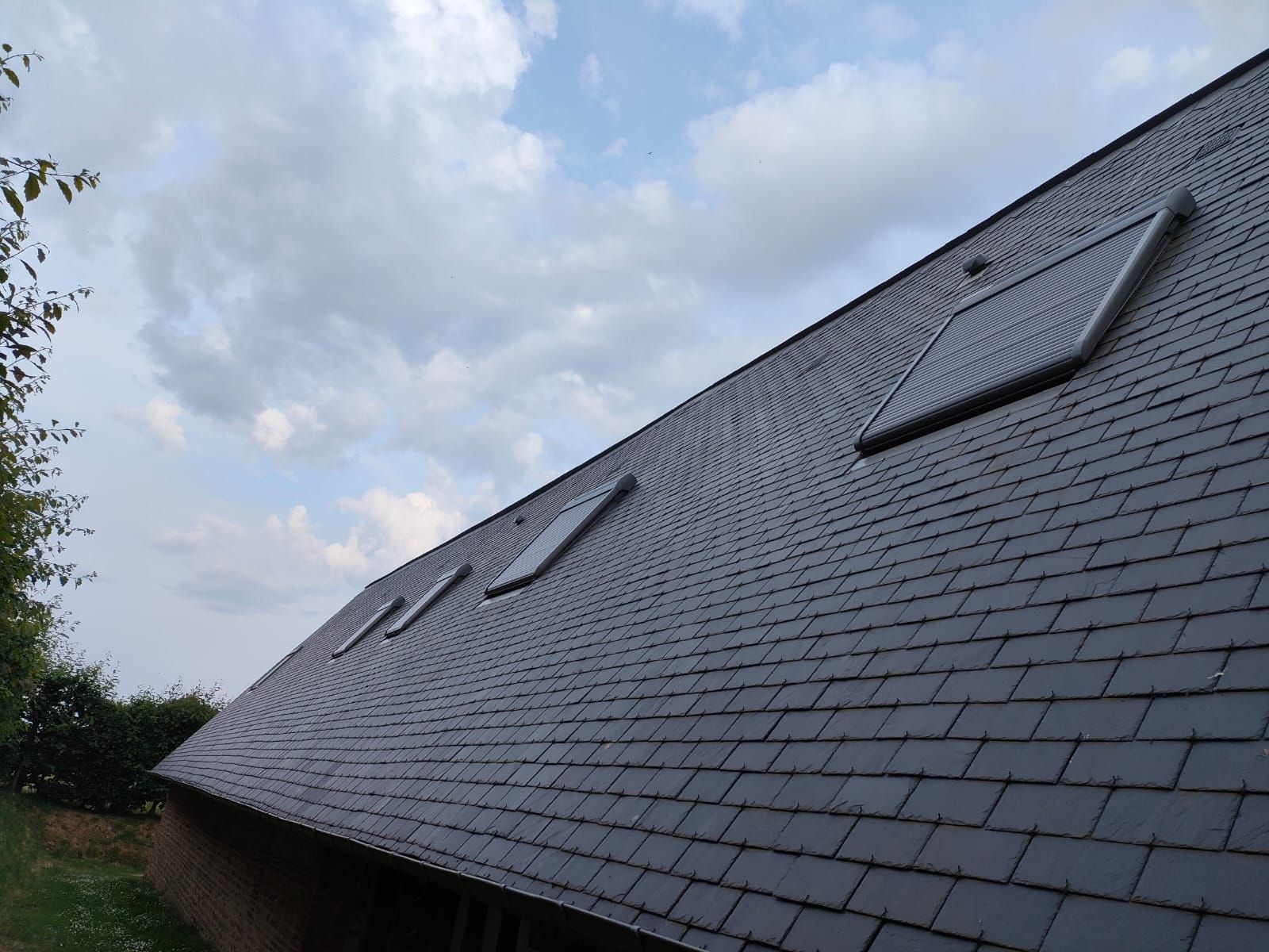 Toit en ardoise percé de trois puits de lumière, sous un ciel nuageux.