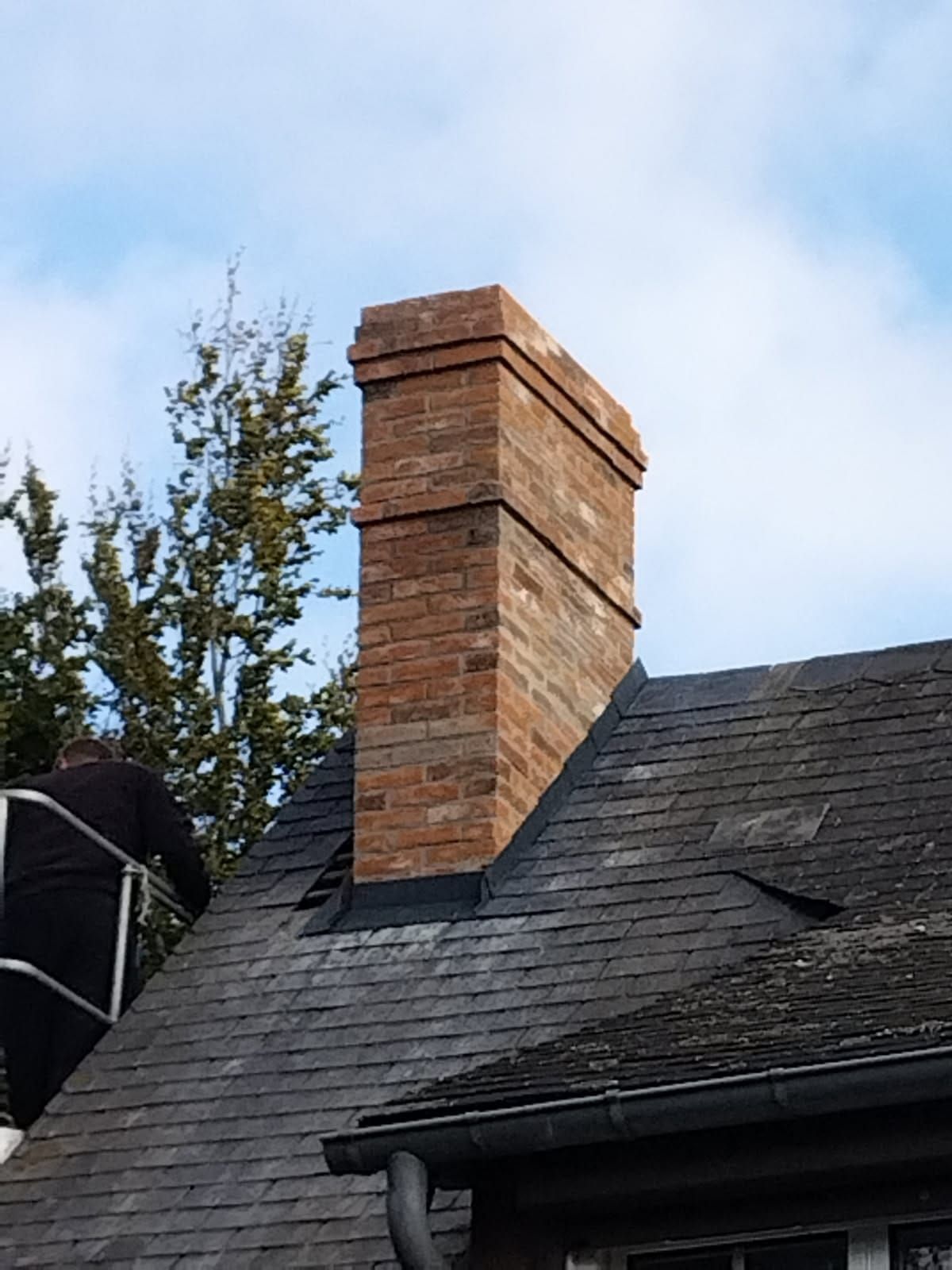 Cheminée en briques sur un toit sombre, avec une personne travaillant sur une échelle. Ciel bleu en arrière-plan.