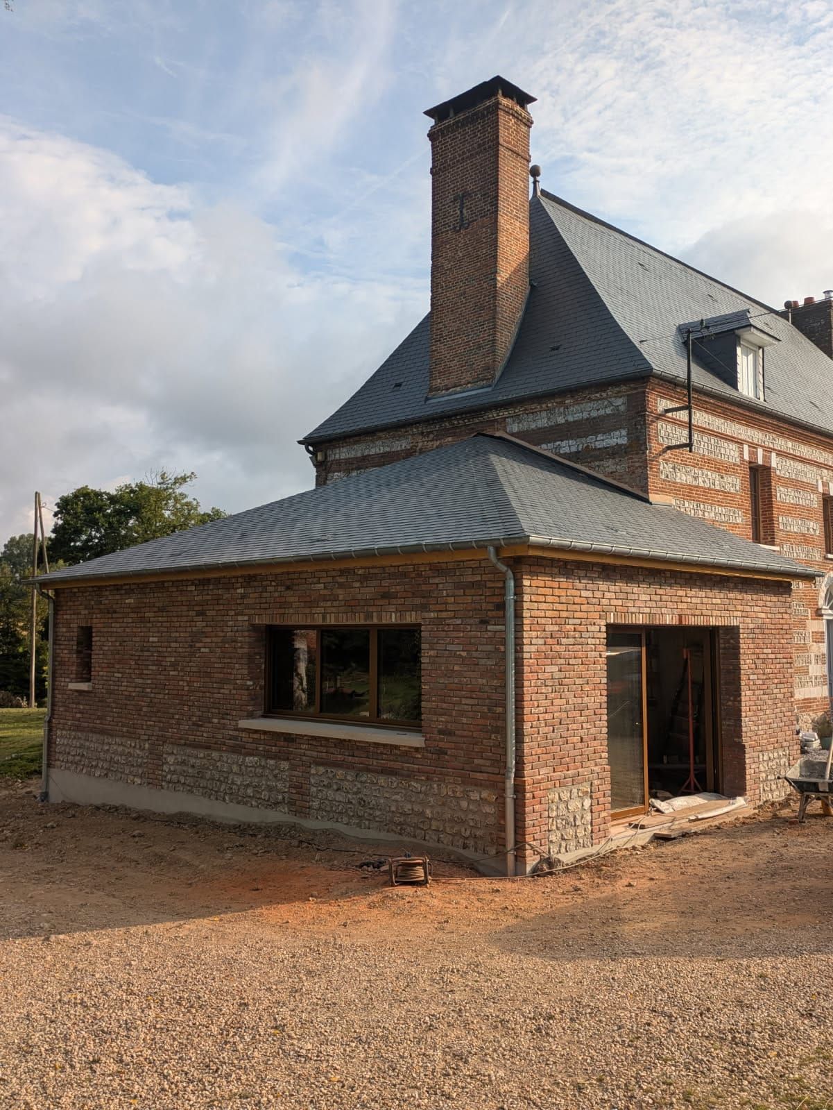 Bâtiment en briques avec une extension neuve en briques, toit gris, haute cheminée en briques et sol en gravier.