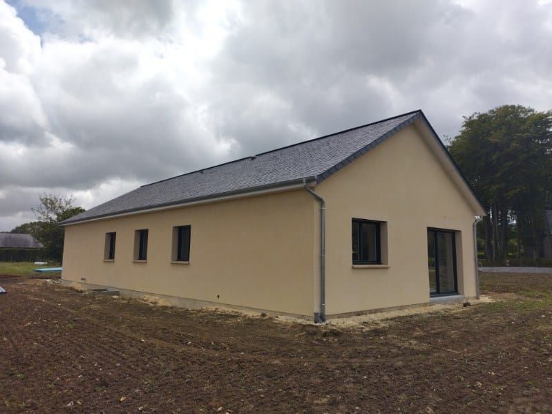 Maison beige au toit, aux fenêtres et à la porte noirs, située dans un champ sous un ciel nuageux.