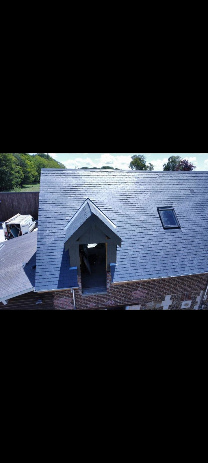 Vue aérienne d'un toit en ardoise avec lucarne et puits de lumière. Arbres verts et route visibles en arrière-plan.