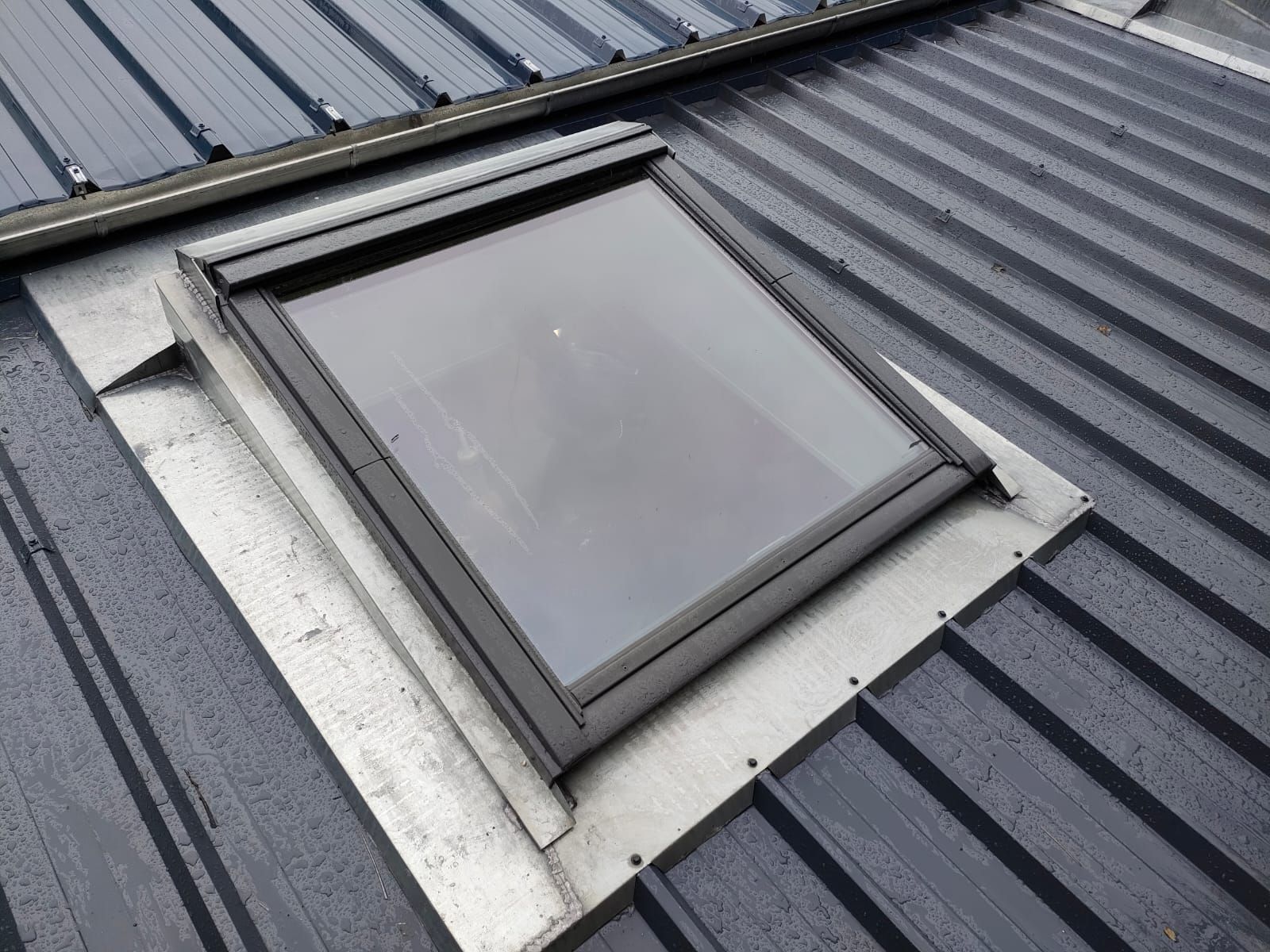 Puits de lumière sur un toit en tôle ondulée foncée. Cadre de fenêtre et solin gris.
