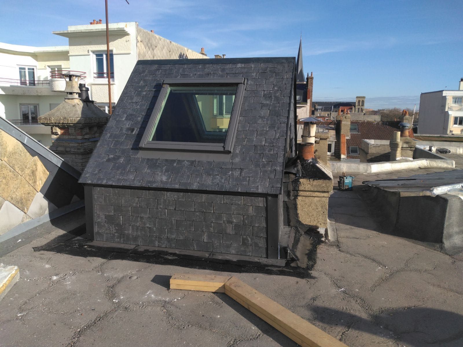 Une lucarne gris foncé avec une fenêtre sur le toit. Une journée ensoleillée, d'autres bâtiments sont visibles.