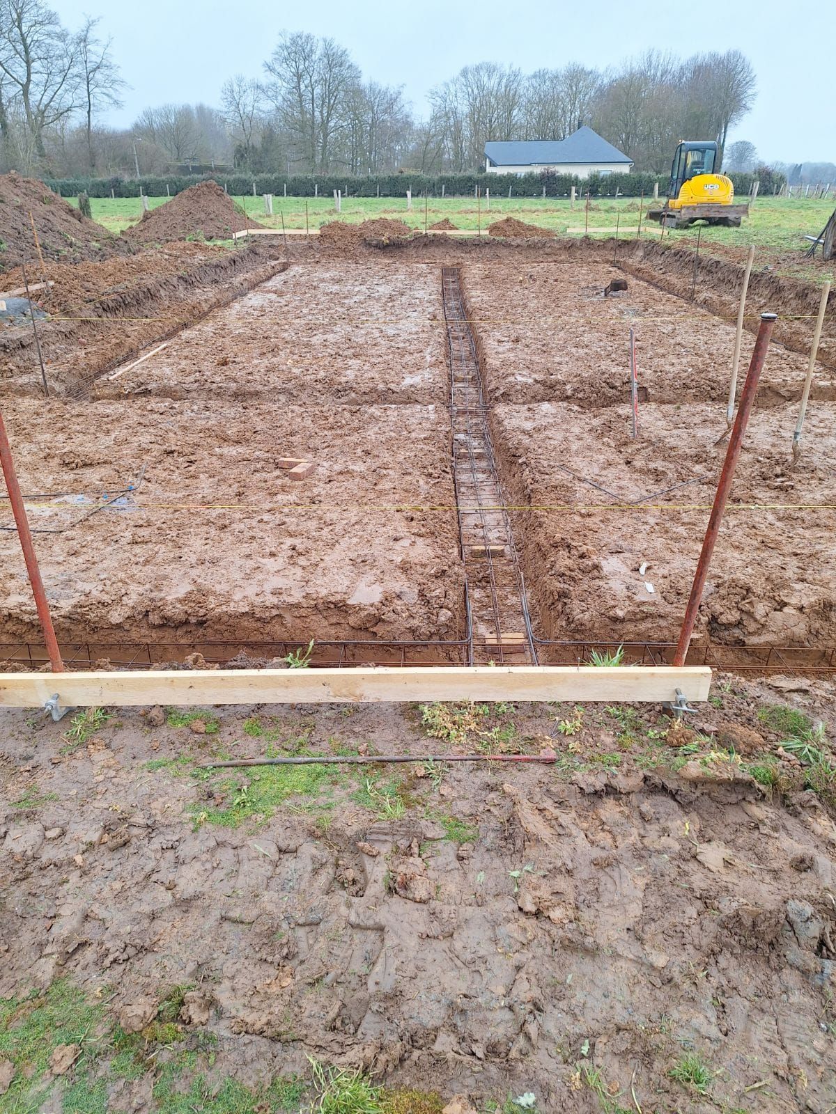 Chantier avec de la boue, des coffrages en bois et des barres d'armature. Une petite pelleteuse est visible au loin.