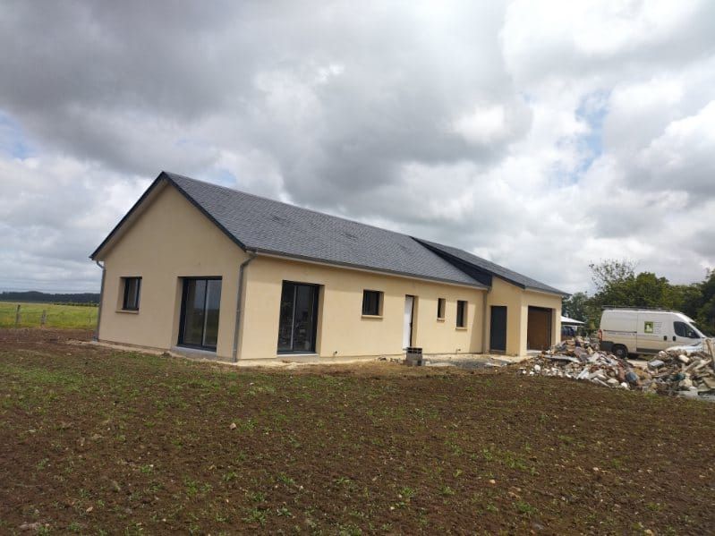 Maison beige au toit sombre et aux encadrements de fenêtres noirs, dans un champ sous un ciel nuageux.