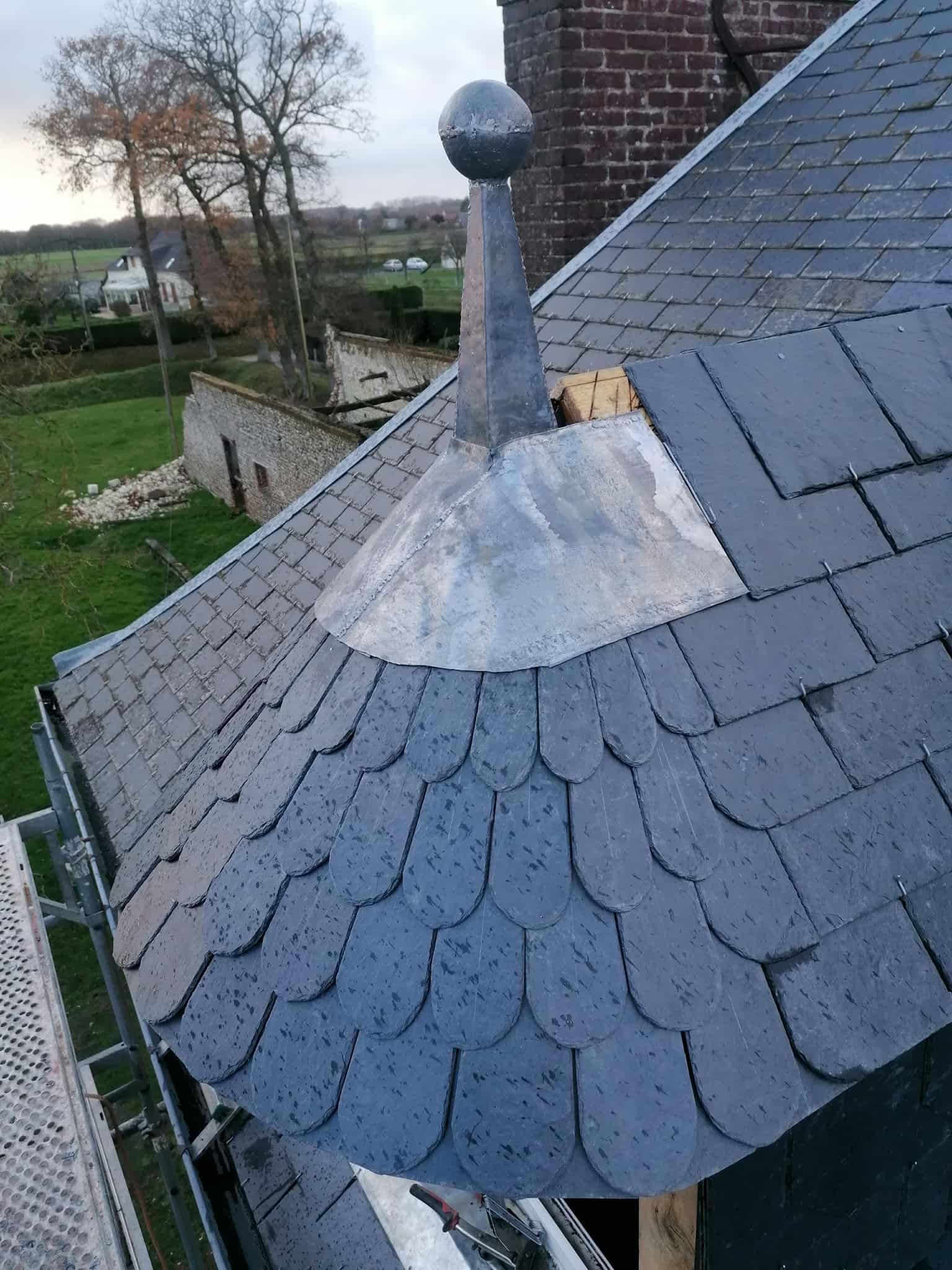 Toit en ardoise orné d'un fleuron et d'une boule, avec un échafaudage partiel au premier plan.