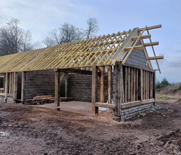 Structure en bois en construction, poutres et chevrons apparents. Extérieur : sol boueux et ciel couvert.