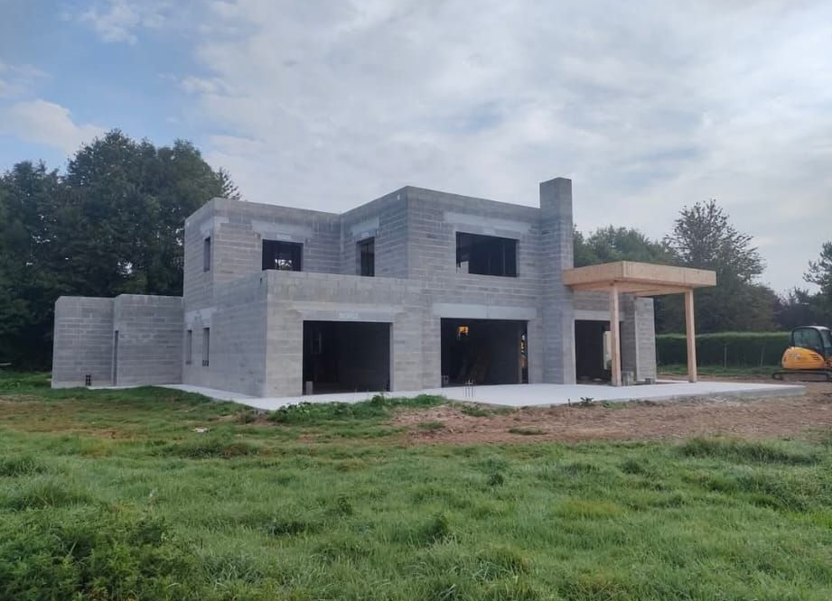 Maison en béton de deux étages en construction, avec de grandes fenêtres et un porche couvert, sur un terrain gazonné.