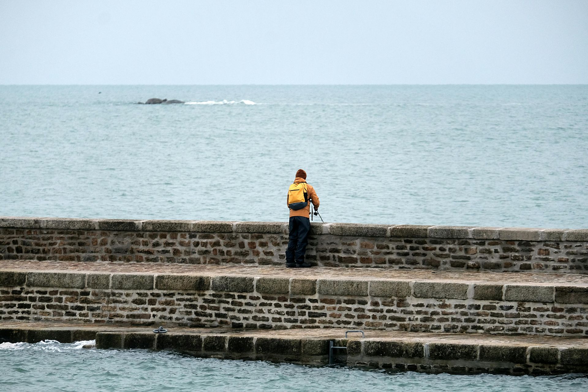 Une personne portant un sac à dos jaune se tient sur une jetée de pierre, regardant l'océan sous un ciel nuageux.